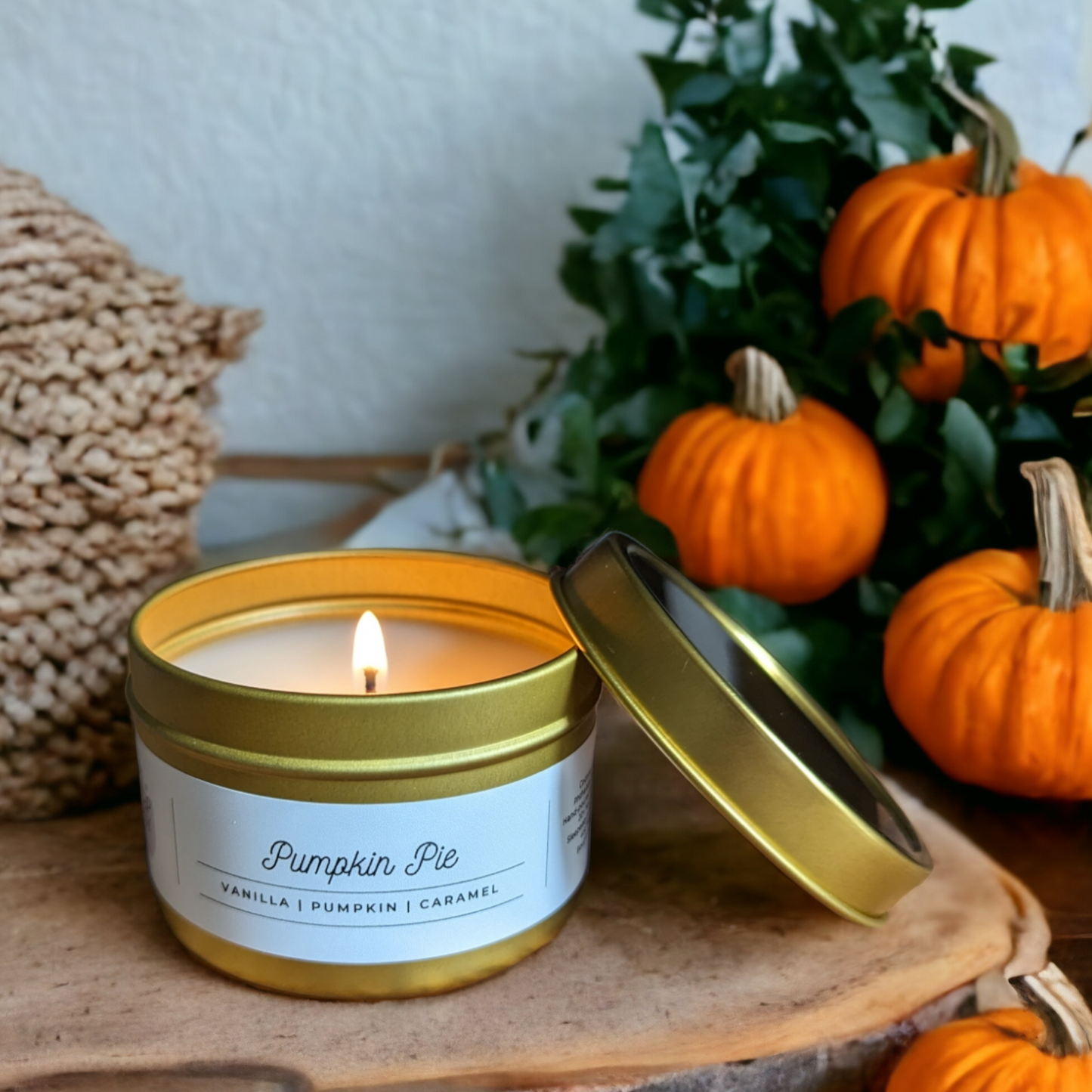 Pumpkin Pie candle. A lit candle in a gold tin sits on a wooden round. Pumpkins and green leaves are in the background.