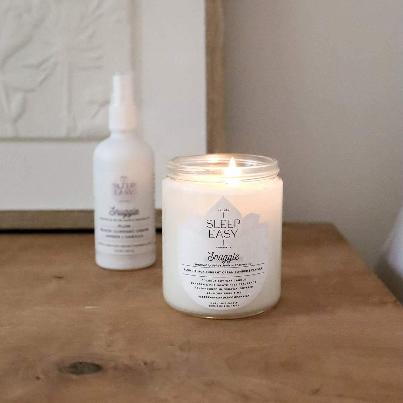 Snuggle coconut soy candle is displayed on top of a wooden table with a wooden picture frame and white picture in the background.