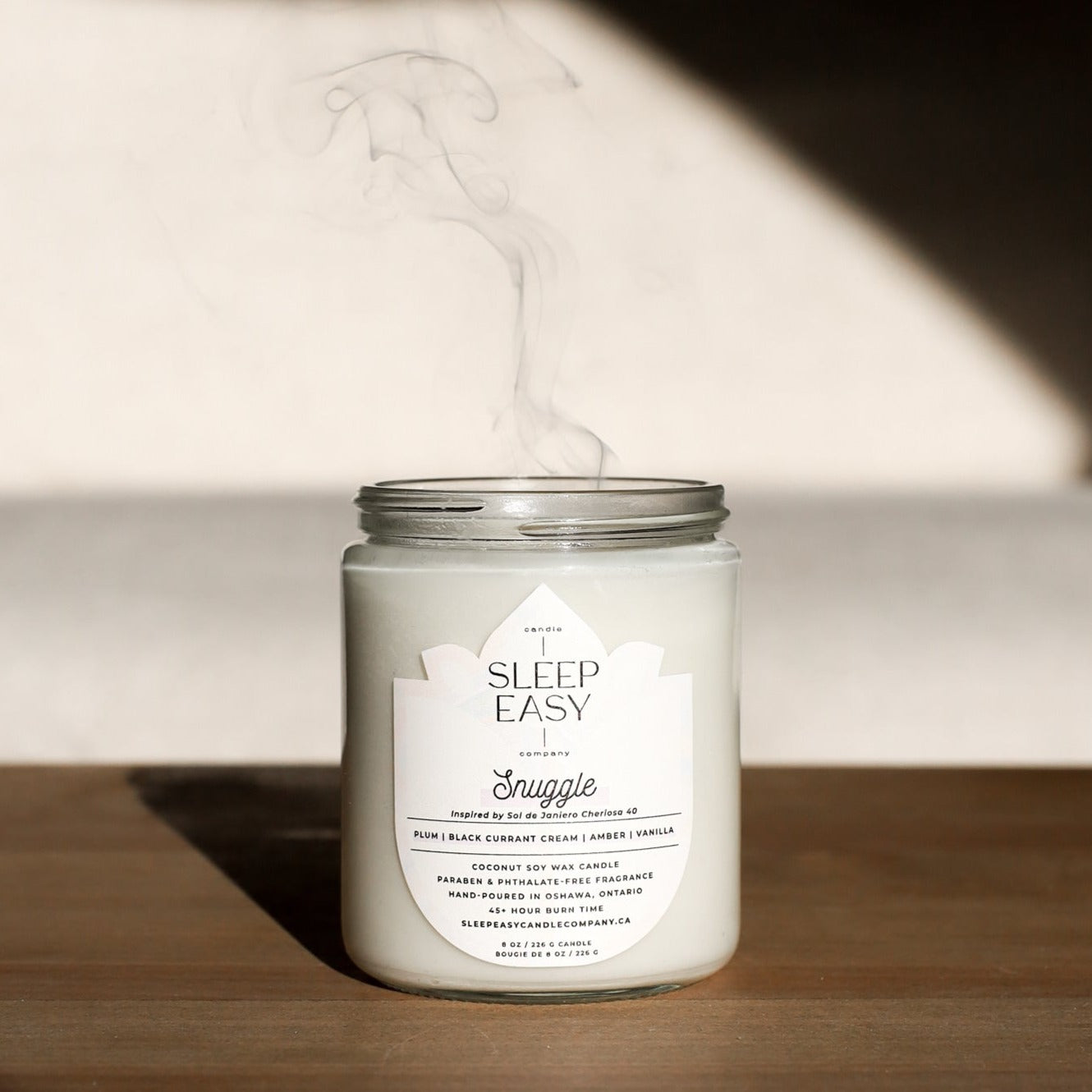 Snuggle Coconut Soy Candle is displayed on a wooden table with shadow lighting. The candle has been blown out and there is smoke above the candle.