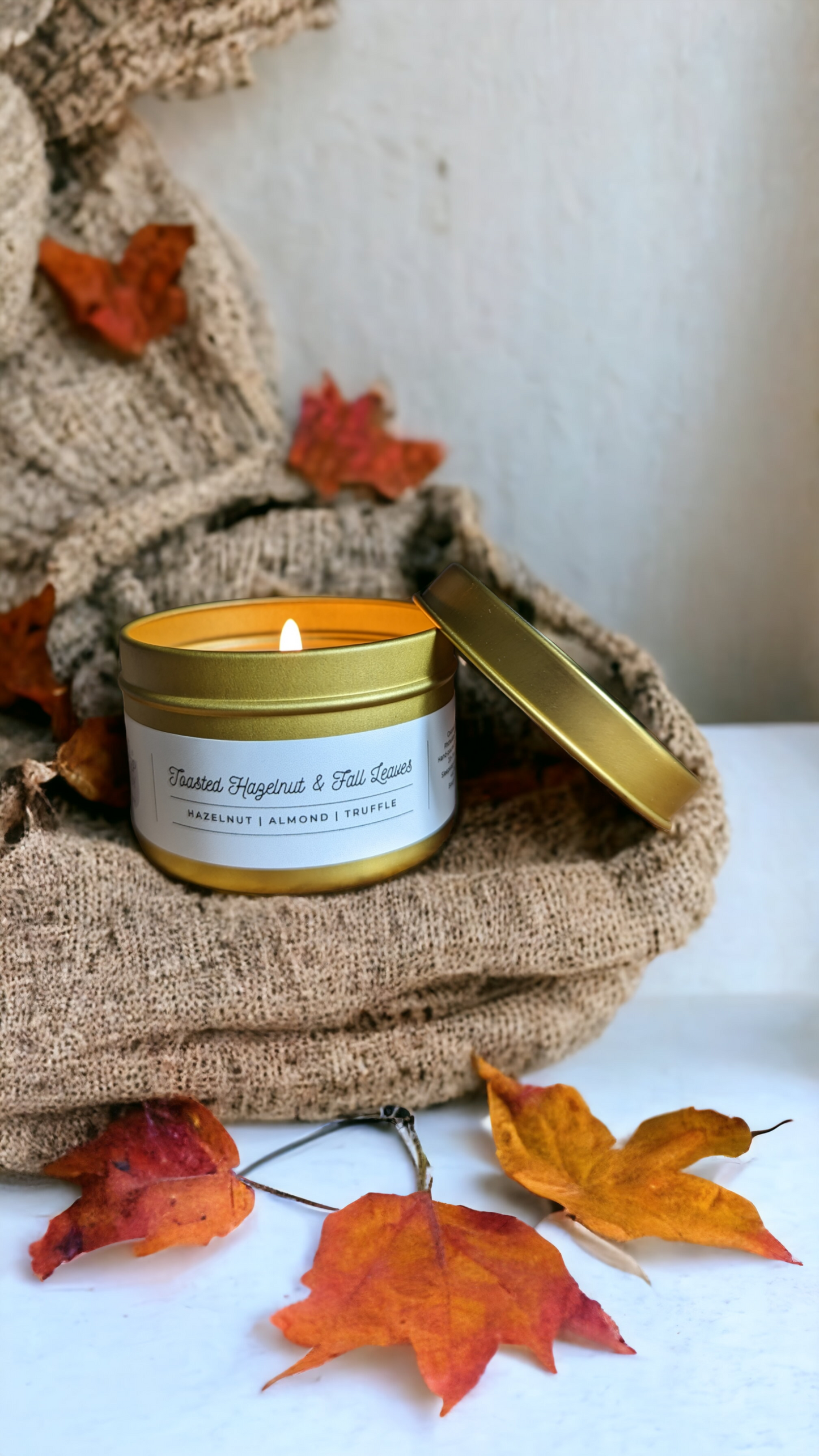 Hazelnut & Fall Leaves Candle in a gold tin jar is displayed on burlap with red fall leaves in background.