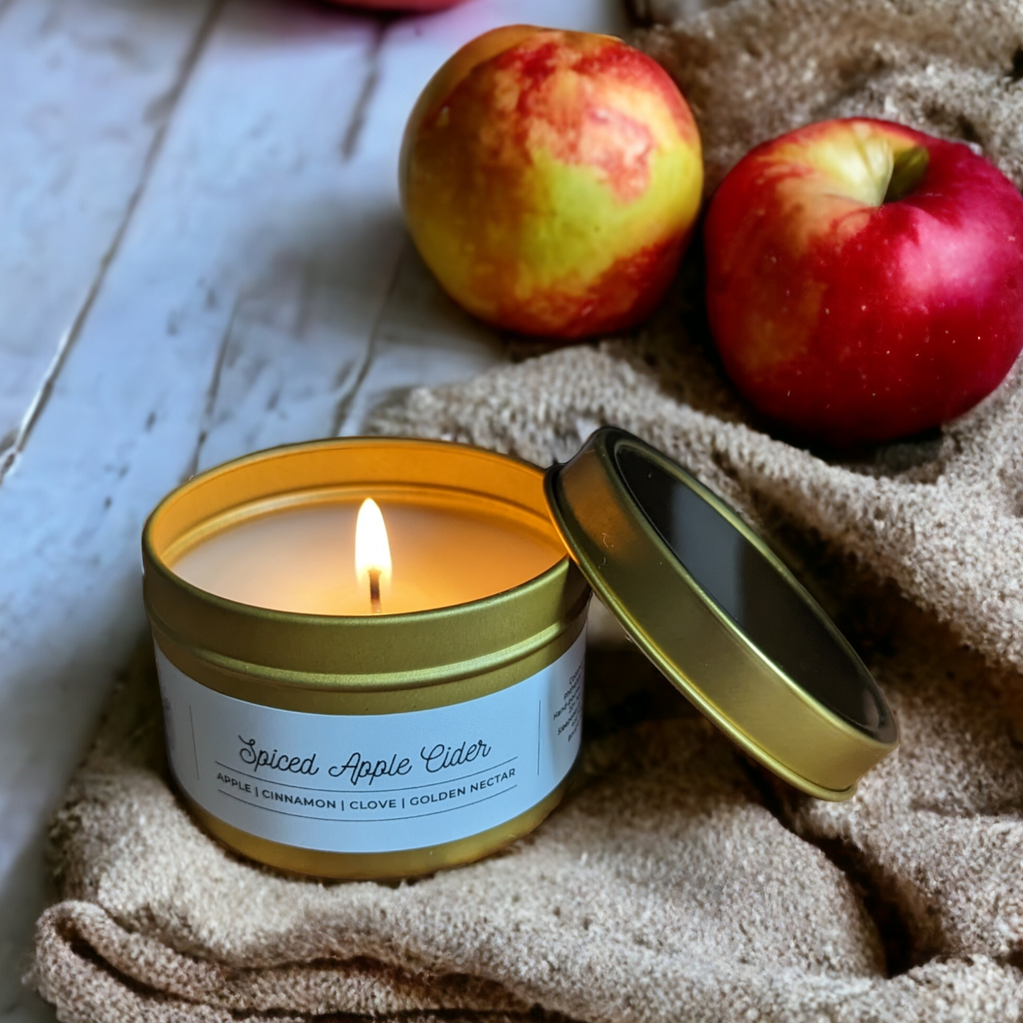 Spiced Apple Cider Candle is displayed on a beige blanket on a white wood floor. Green and red apples lay in the background.