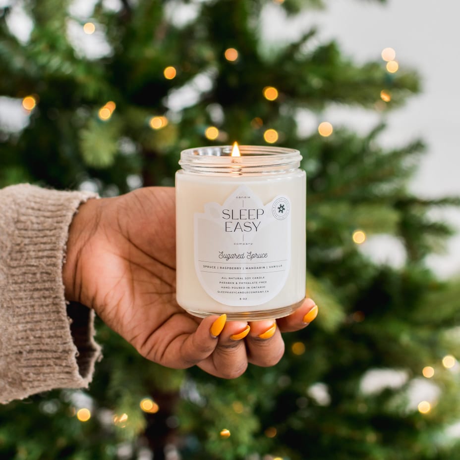 All natural eco friendly soy candle is being held in a woman's hand in front of a green lit Christmas tree. The candle label reads, sugared spruce soy candle, by sleep easy candle company.