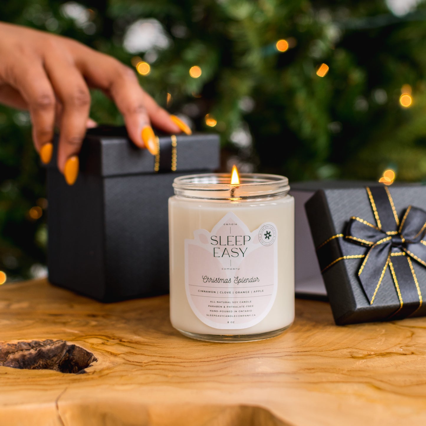 White soy candle on wood table infront of green Christmas tree with white lights. A black gift box with a black bow and gold trim is behind the candle.