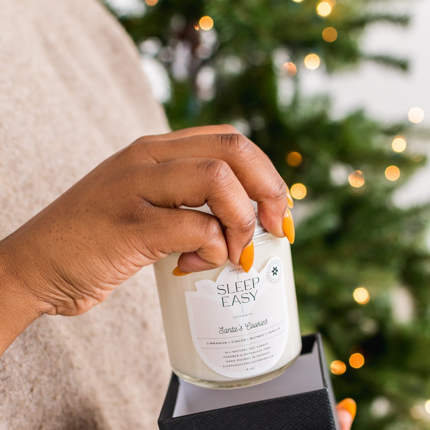 Eco friendly soy candle is being lifted out of a gift box in front of a christmas tree. The Christmas tree is lit with Christmas lights. 