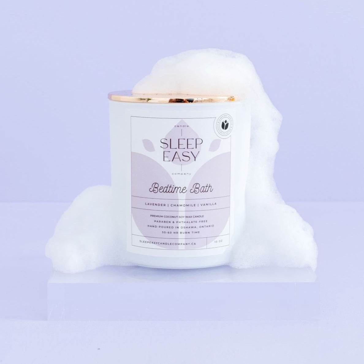 Bedtime Bath coconut soy candle, white candle jar with gold lid is displayed on a purple background. The candle has bath bubbles on the lid and sides of candle. The label is white and light blue. Logo in the background of the label looks like a flower. Hand poured coconut soy candles. Handmade in Oshawa, Ontario. Luxury candles Canada. Scented candles Canada. Luxury Scented candles. Free Canadian shipping on orders over $99.99.