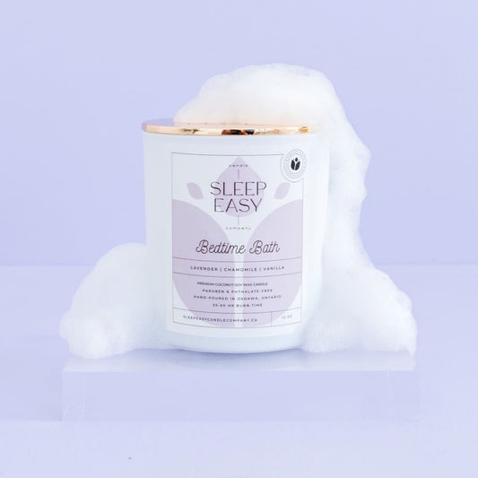 Bedtime Bath coconut soy candle, white candle jar with gold lid is displayed on a purple background. The candle has bath bubbles on the lid and sides of candle. The label is white and light blue. Logo in the background of the label looks like a flower. Hand poured coconut soy candles. Handmade in Oshawa, Ontario. Luxury candles Canada. Scented candles Canada. Luxury Scented candles. Free Canadian shipping on orders over $99.99.