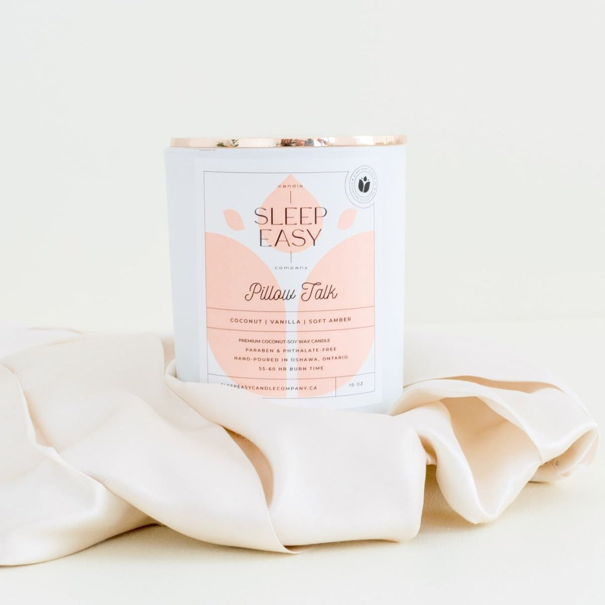 Pillow Talk coconut soy candle. Coconut, vanilla, soft amber fragrance. white candle jar with gold lid is displayed on an elegant beige background. The candle is in the middle of beige silk sheets. The label is white and light peach colour. Logo in the background of the label looks like a delicate flower. Handmade in Oshawa, Ontario. Luxury candles Canada. Scented candles Canada. Luxury Scented candles. Free Canadian shipping on orders over $99.99. romantic scents for the bedroom. sexy scents.