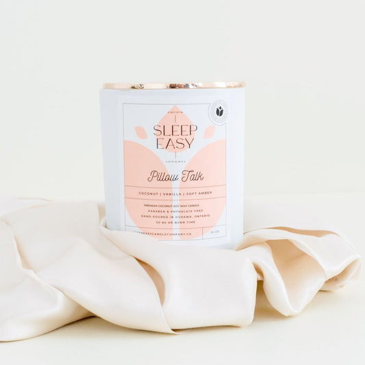 Pillow Talk coconut soy candle. Coconut, vanilla, soft amber fragrance. white candle jar with gold lid is displayed on an elegant beige background. The candle is in the middle of beige silk sheets. The label is white and light peach colour. Logo in the background of the label looks like a delicate flower. Handmade in Oshawa, Ontario. Luxury candles Canada. Scented candles Canada. Luxury Scented candles. Free Canadian shipping on orders over $99.99. romantic scents for the bedroom. sexy scents.