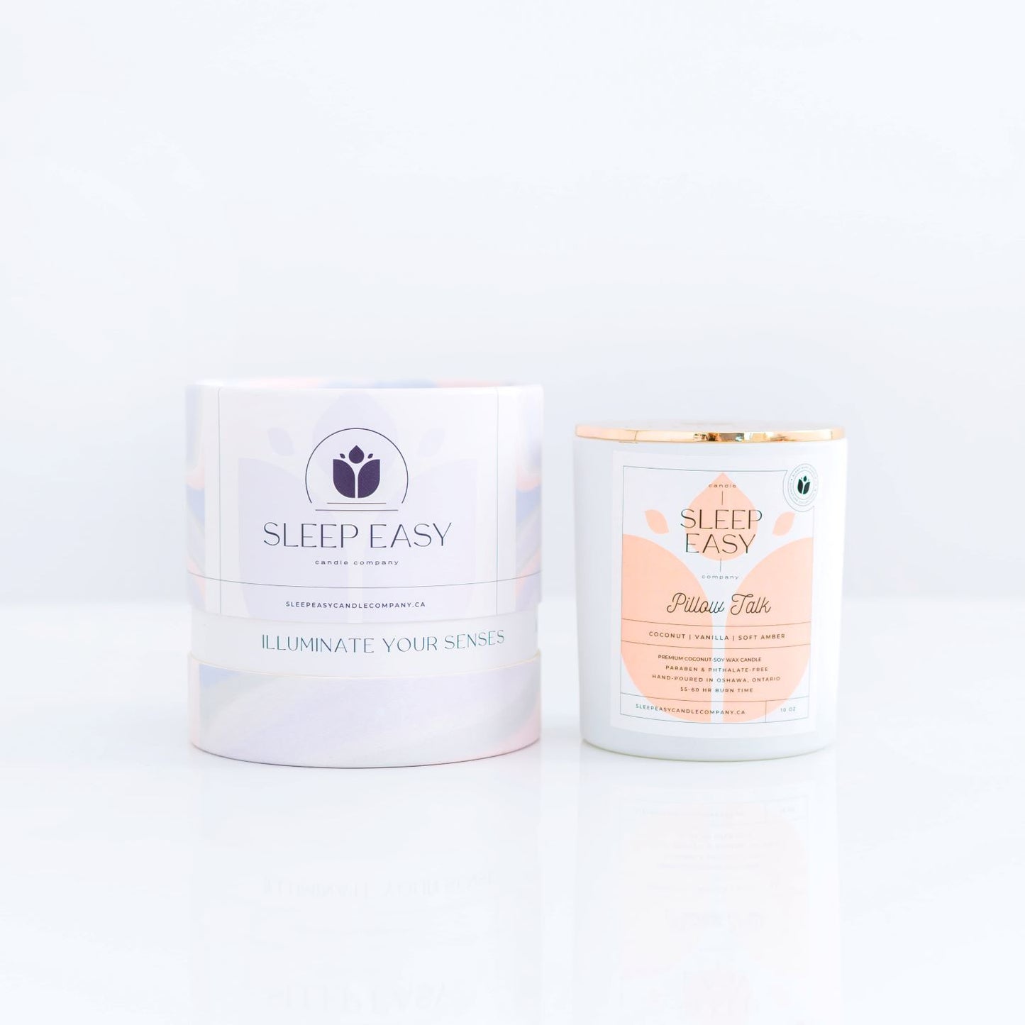Pillow Talk coconut soy candle, white candle jar with gold lid is displayed on a white background. The label is white and light peach. Logo in the background of the label looks like a delicate flower. Beside the candle jar is a branded tube box with a wavy multi colour texture. Hand poured coconut soy candles. Handmade in Oshawa, Ontario. Luxury candles Canada. Scented candles Canada. Luxury Scented candles. Luxury soy coconut candles Canada. Free Canadian shipping on orders over $99.99.