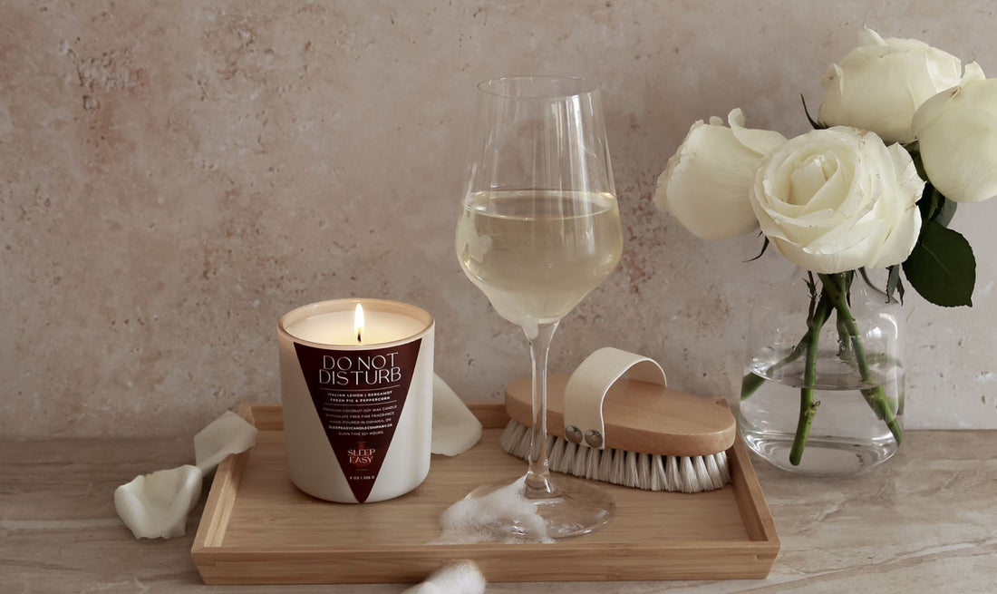 Our Do Not Disturb candle is placed on a bath tray with a wine glass filled with white wine. Bubbles from the bath are splatter across the bath tray. White roses are placed in a vase behind the candle. 