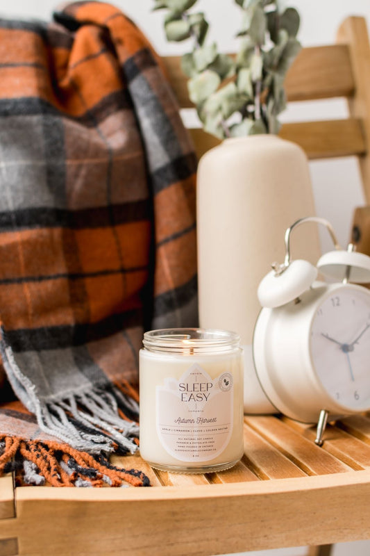 A candle named autumn harvest is placed on a wooden chair. A orange black and grey flannel blanket lays in the background. 