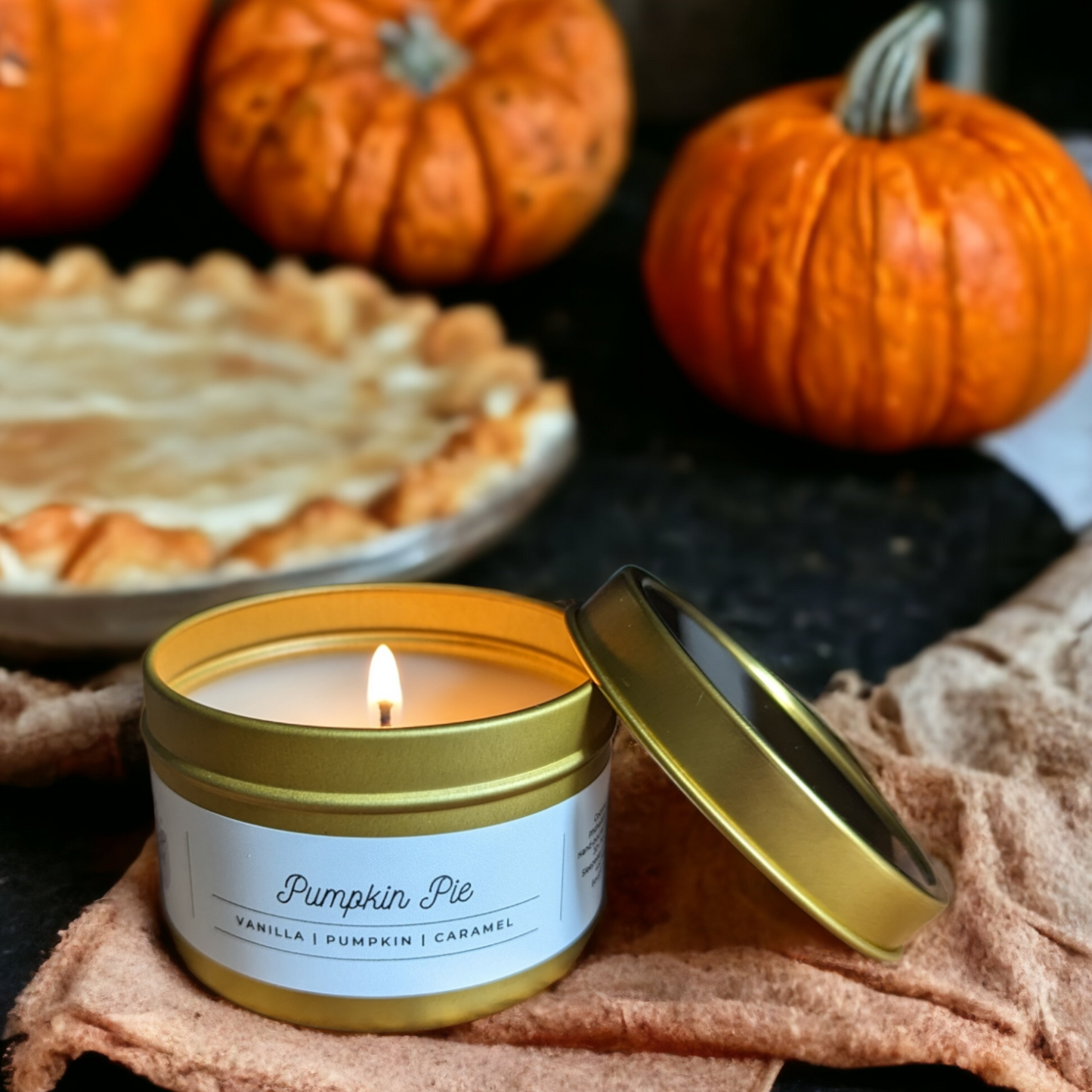 A lit candle in a 4oz gold tin jar sits on top of a blanket. Pumpkins and pumpkin pie is in the background.Pumpkin Pie Candle, Fall Candle, Fall Scented Candles.