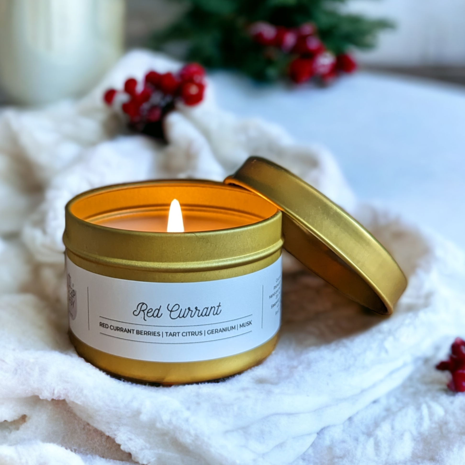 4oz gold travel tin candle displayed on a white blanket in front of red berries and pine bush. Red currant coocnut soy candle is inspired by Vovito Red Currant home fragrance. 