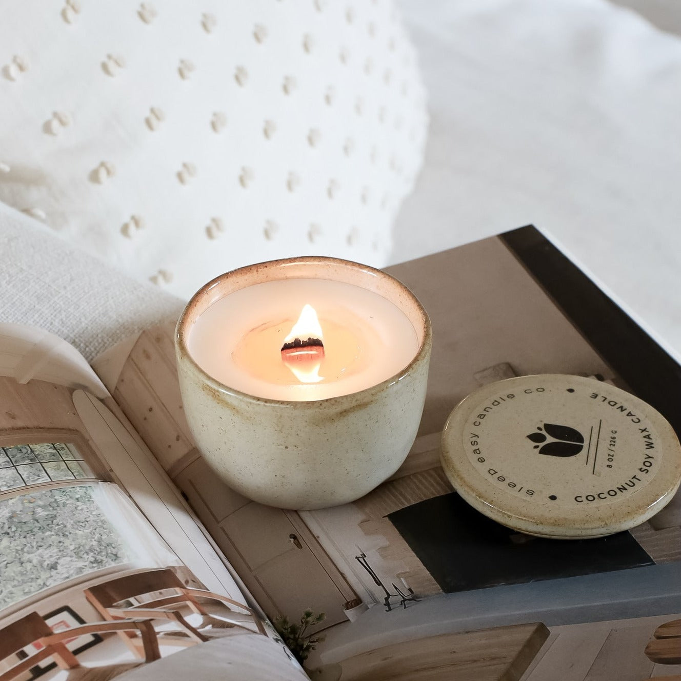 Brown Ceramic Candle on top of a book and white sheets on a bed. 
