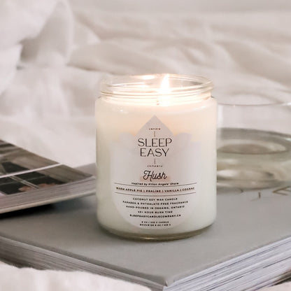 Hush coconut soy candle is displayed on top of a grey book with a white glass with white wine in the background. On top of a white linen sheets. 