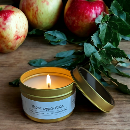 Spiced Apple Cider Candle in a gold tin sits on a wooden table with green leaves and green and red apples in background.