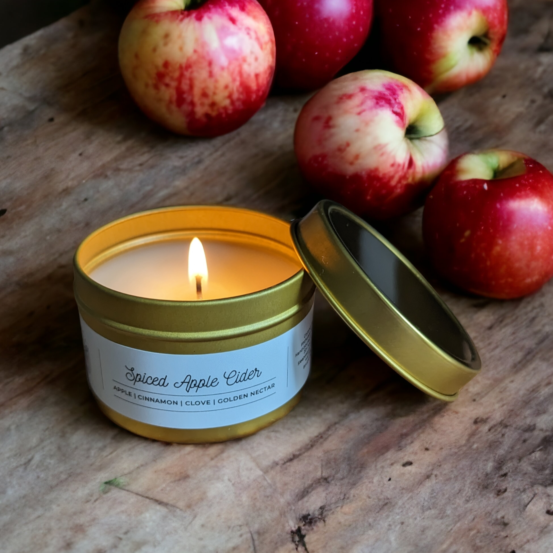 Spiced Apple Cider Candle in a Gold Tin is displayed on a wooden table with green and red apples in background. 