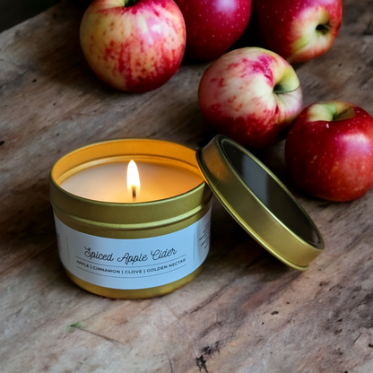 Spiced Apple Cider Candle in a Gold Tin is displayed on a wooden table with green and red apples in background. 