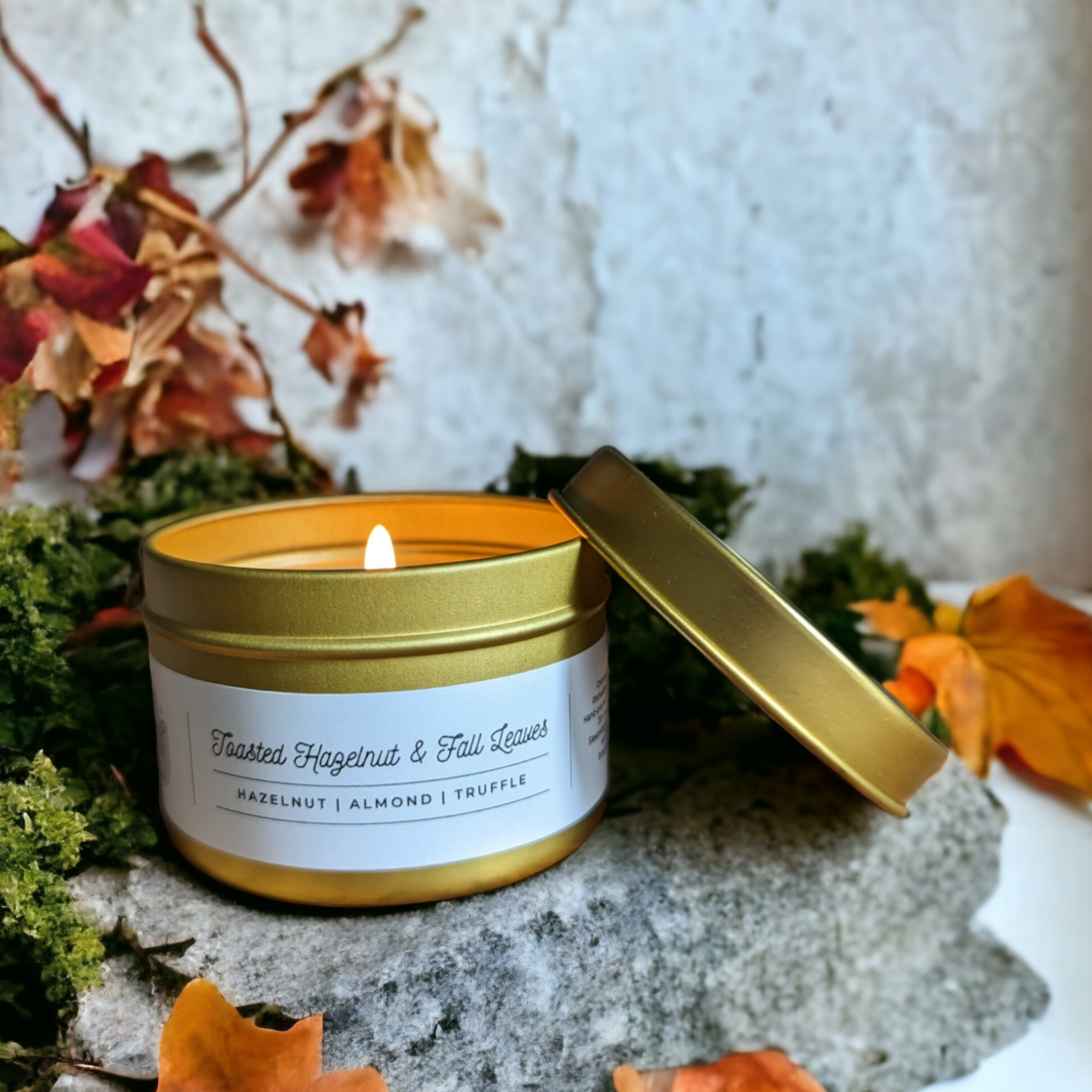 Toasted Hazelnut & Fall Leaves Candle in a gold tin, is displayed on a piece of stone. With orange & red fall leaves and green moss in background.