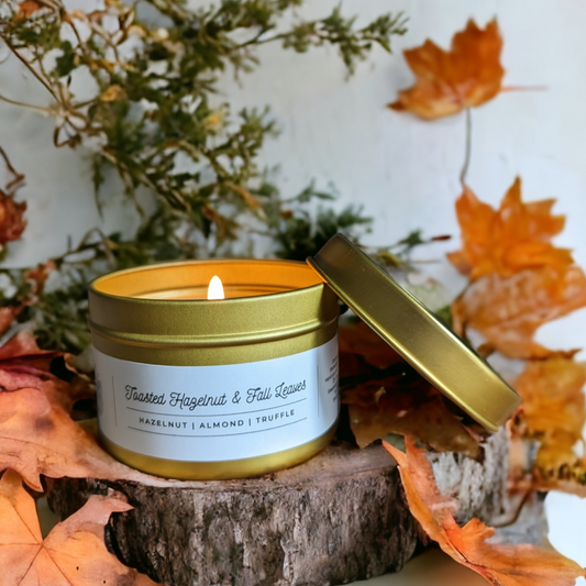 Toasted Hazelnut & Fall Leaves Gold Tin Candle is displayed on a wood round, with green branches and orange fall leaves in background. 