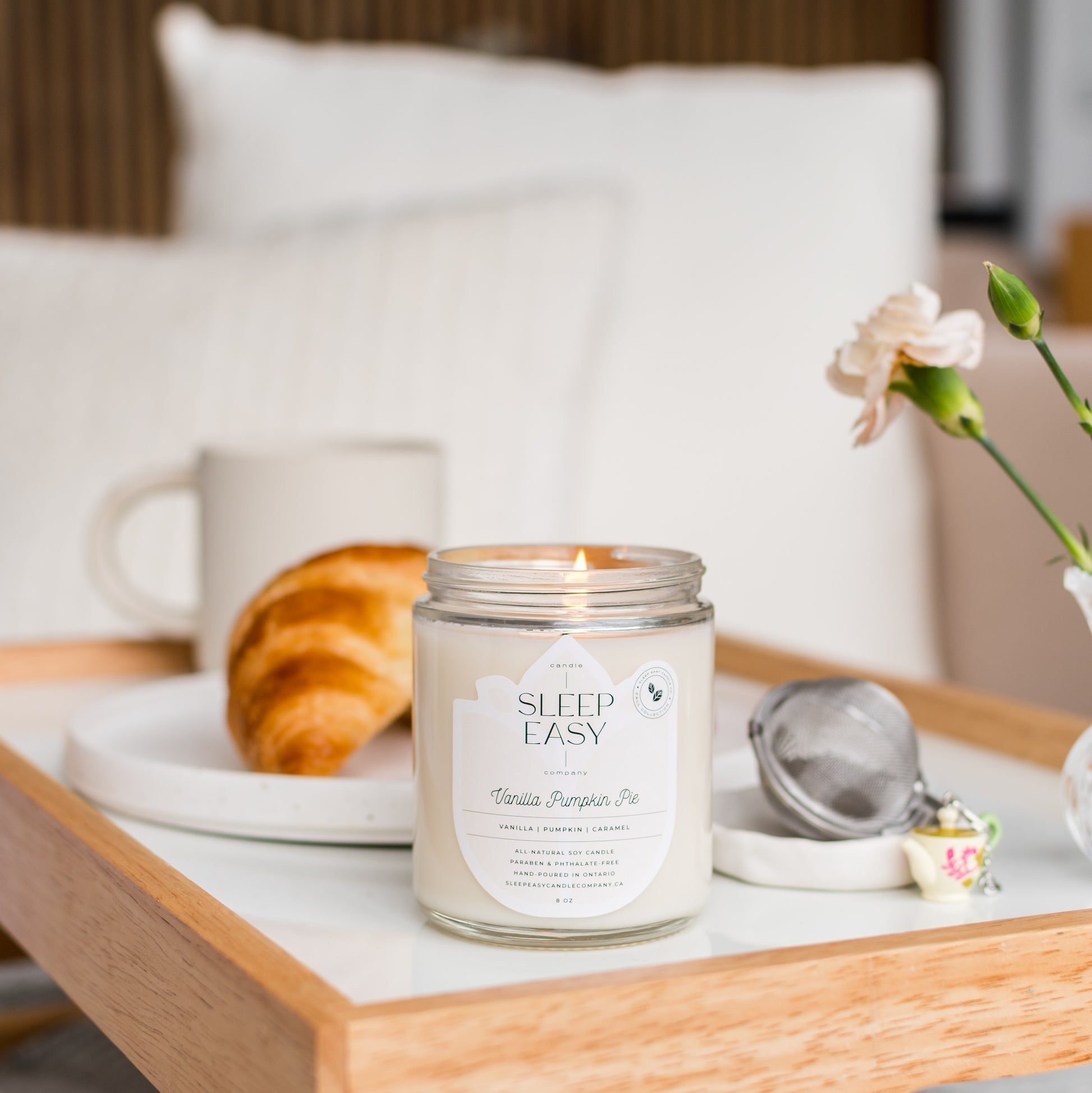 Vanilla Pumpkin Pie Candles is displayed on a white serving tray, with a croissant and tea on the side, the serving tray is displayed on a bed with linen sheets and pillows 