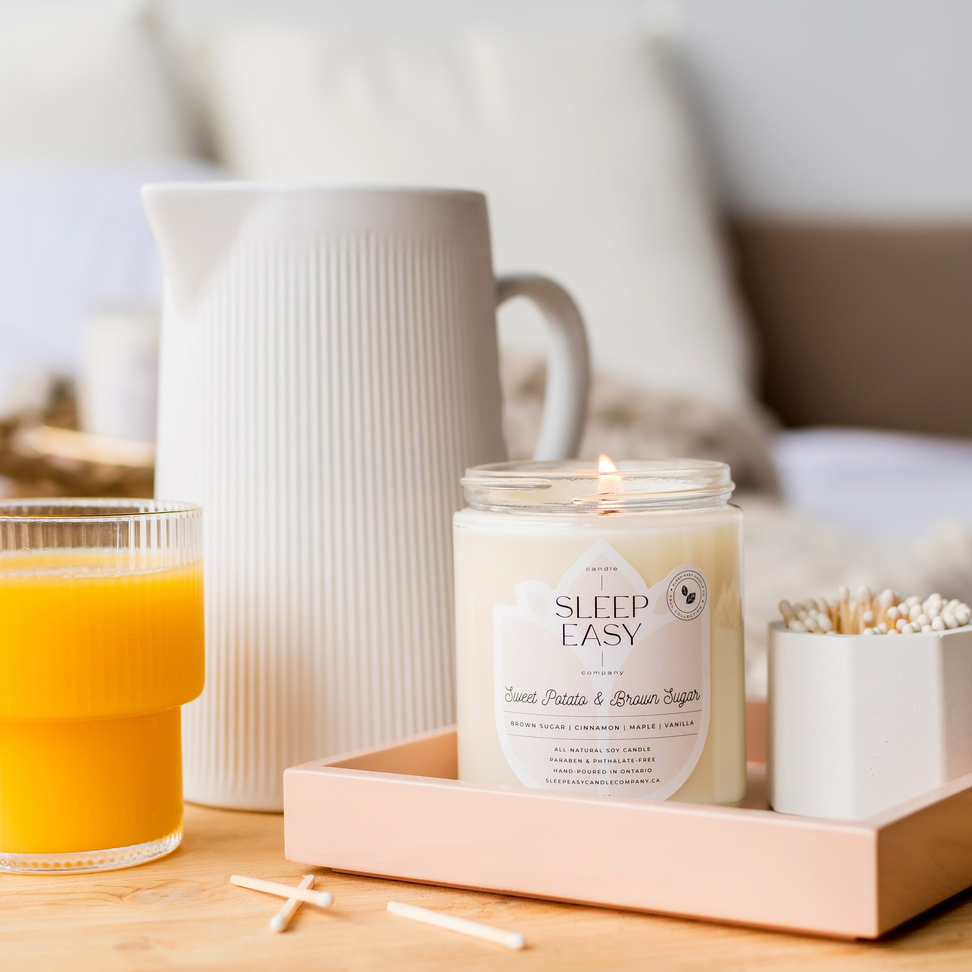 All natural soy candle is displayed on a pink tray with white matches on the side. The tray is on a bed of linen sheets and pillows. Orange juice is on the side of the tray. 