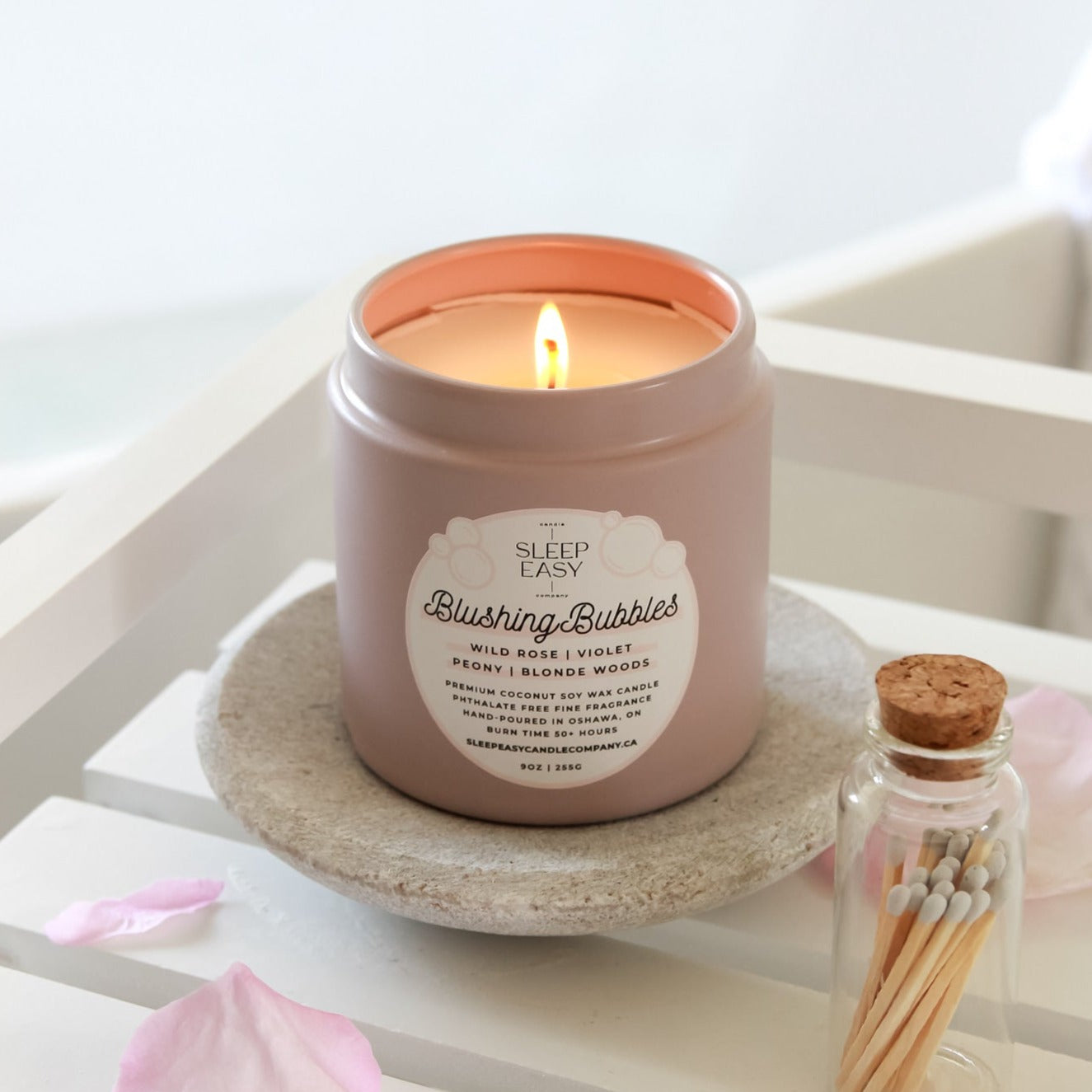 Blushing Bubbles candle is displayed in front of a bathtub filled with water and rose petals. The candle sits on a stone display with white matches and flower petals on the side.