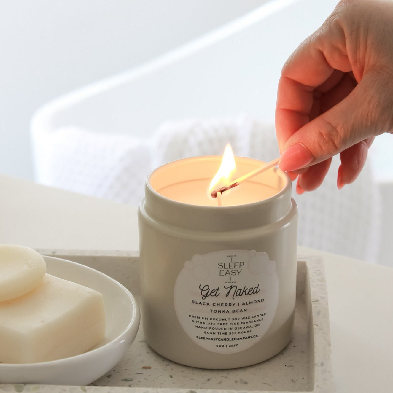 Get Naked Coconut Soy Candle is displayed in a bathroom on a ceramic dish. A woman is lighting the candle with a matchstick. 