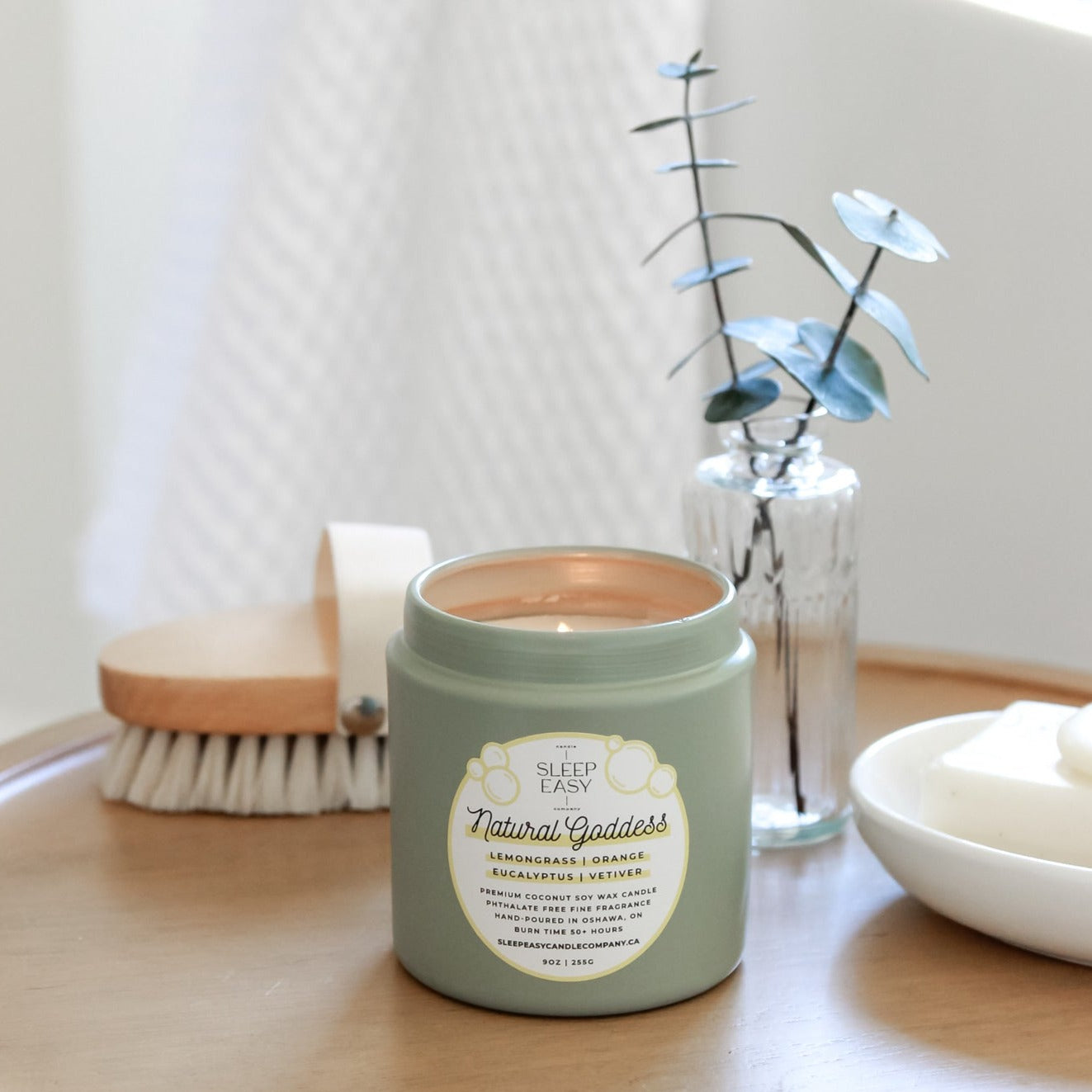 Natural Goddess Coconut Soy Candle is made with Lemongrass, Eucalyptus, and Orange Fragrance. The candle is in a green tin jar, displayed on a wooden table. A soap dish with natural soap is in the background as well as a vase with two eucalyptus stems. 