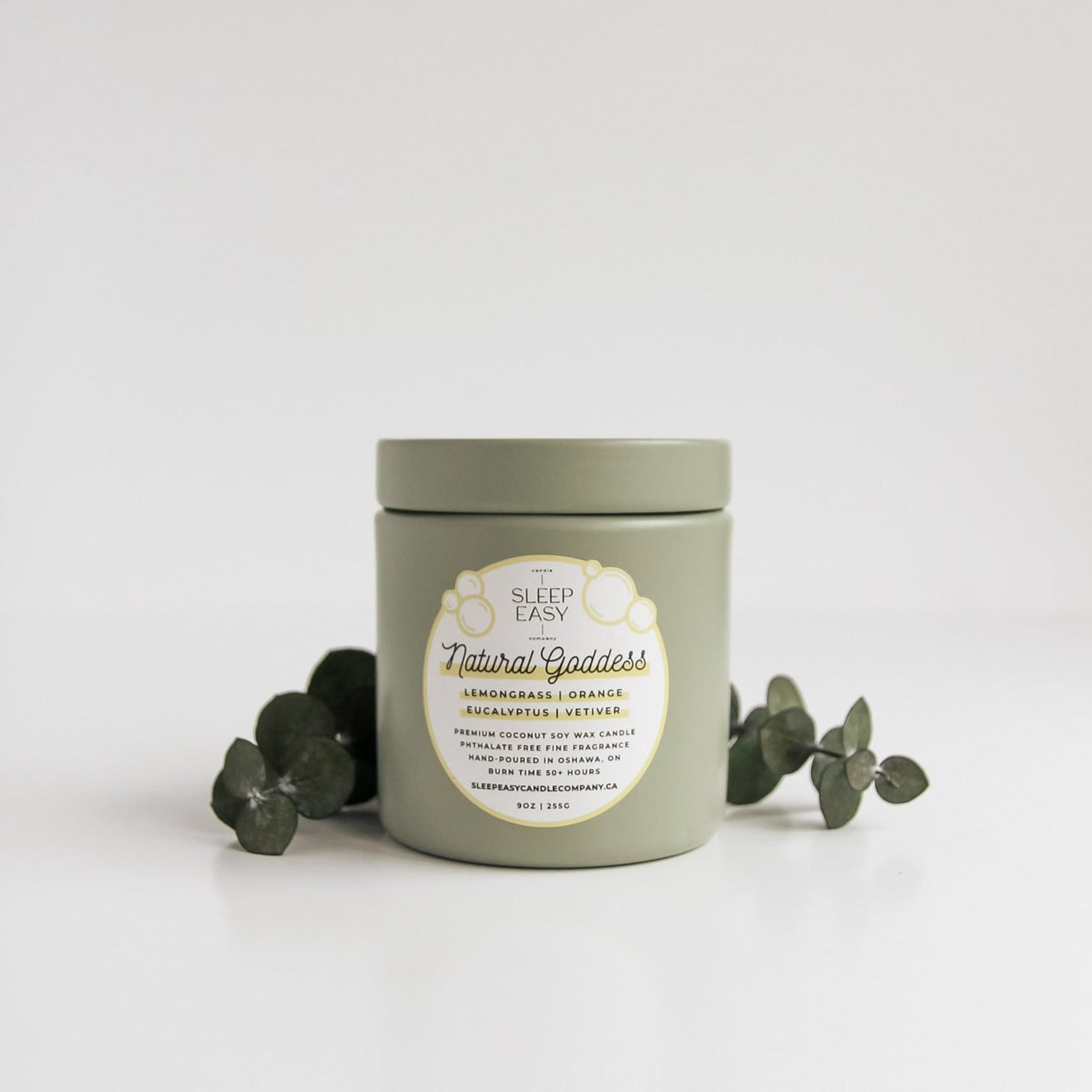 Natural Goddess Coconut Soy Candle is displayed on a white background with eucalyptus stems in the background. 